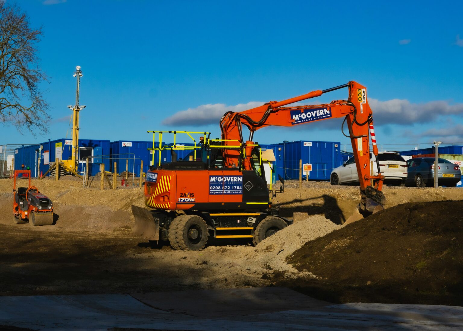 An orange excavator on a construction site. A smaller orange vehicle is to the left.