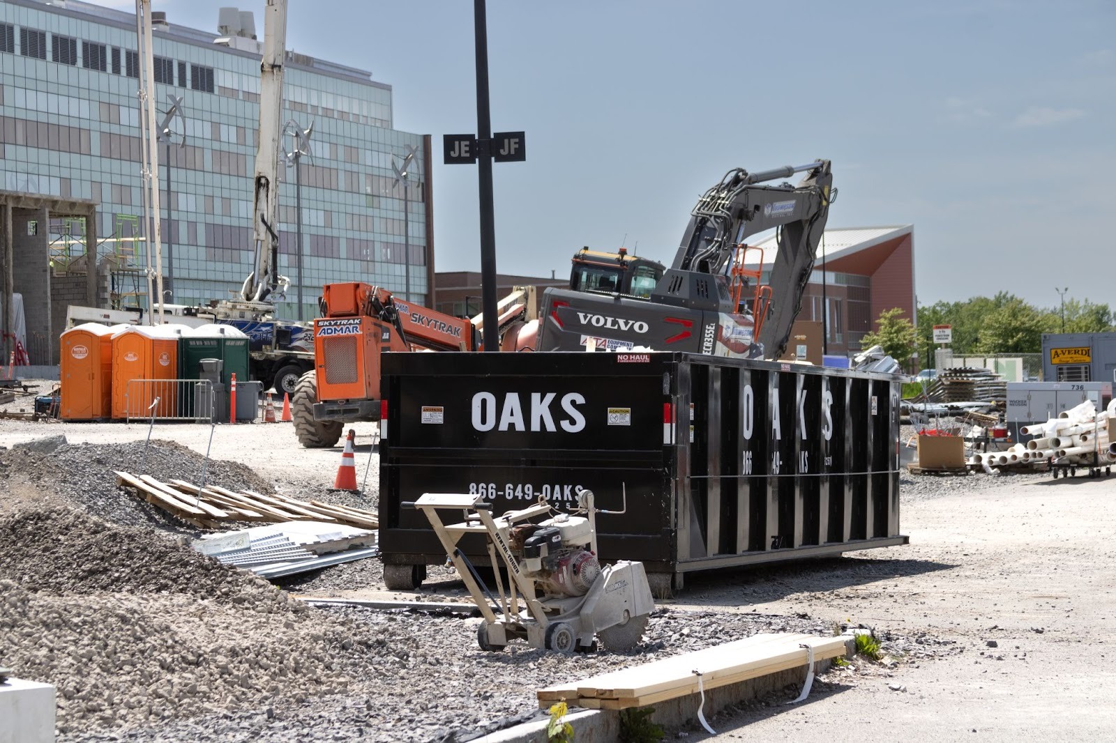 A construction site with a black dumpster and an excavator. The dumpster has 'OAKS' written on it.