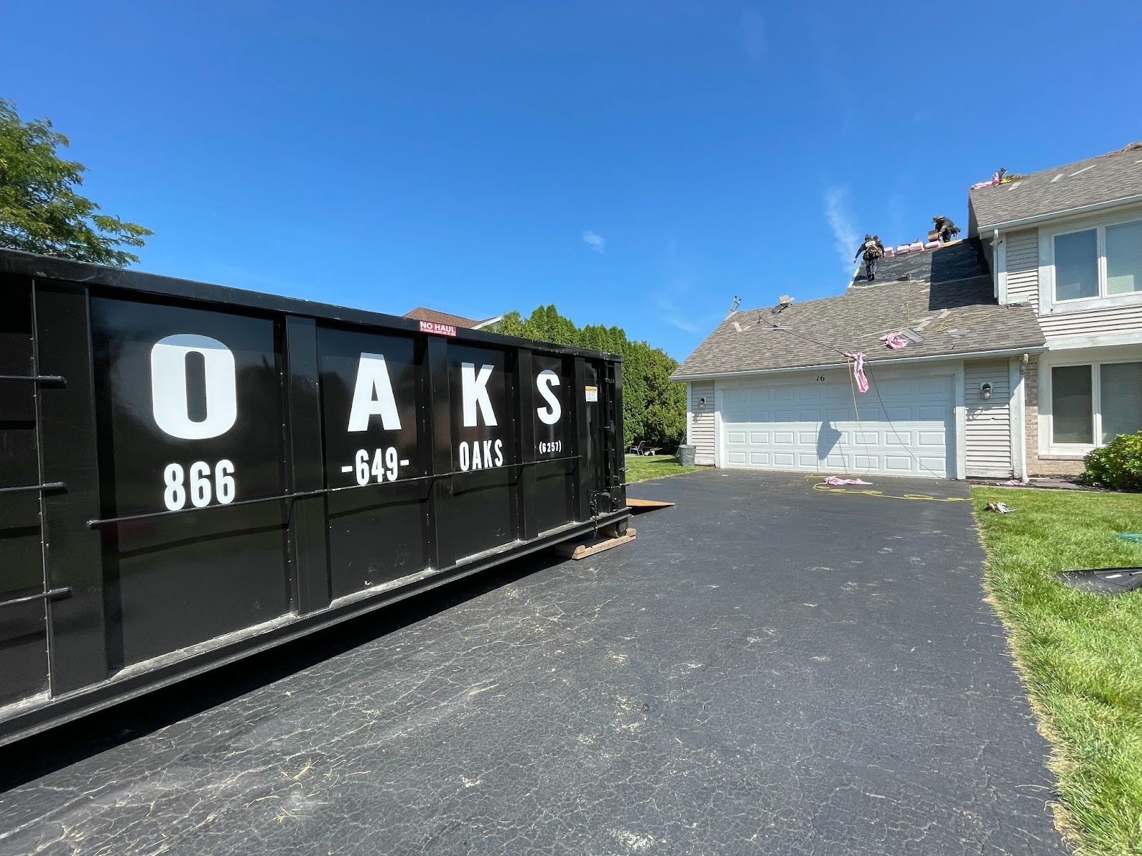 a large black dumpster on a driveway. The dumpster has O-A-K-S and a phone number on it.