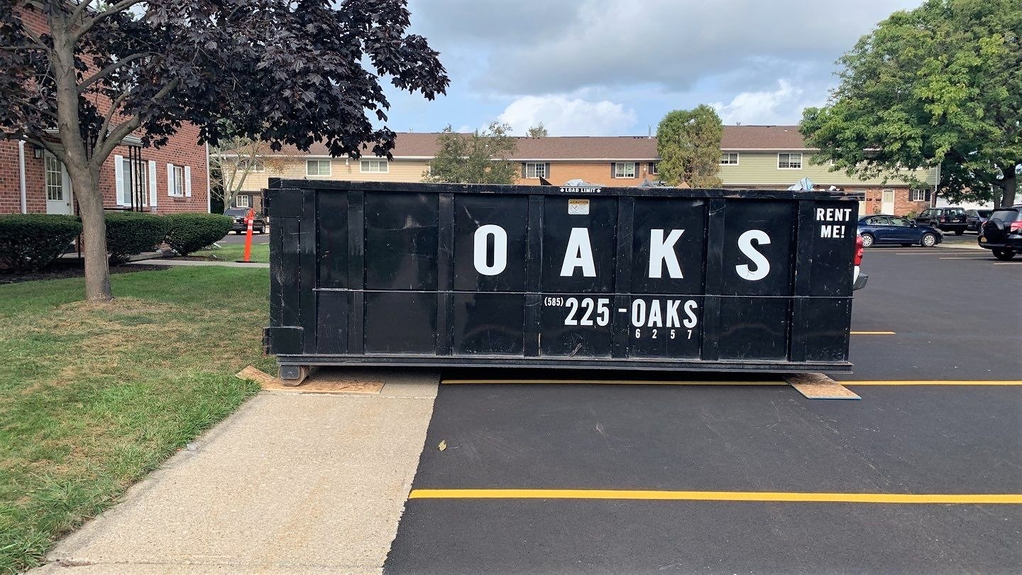 A large black dumpster with 'OAKS' written on it. The dumpster is on a parking lot.