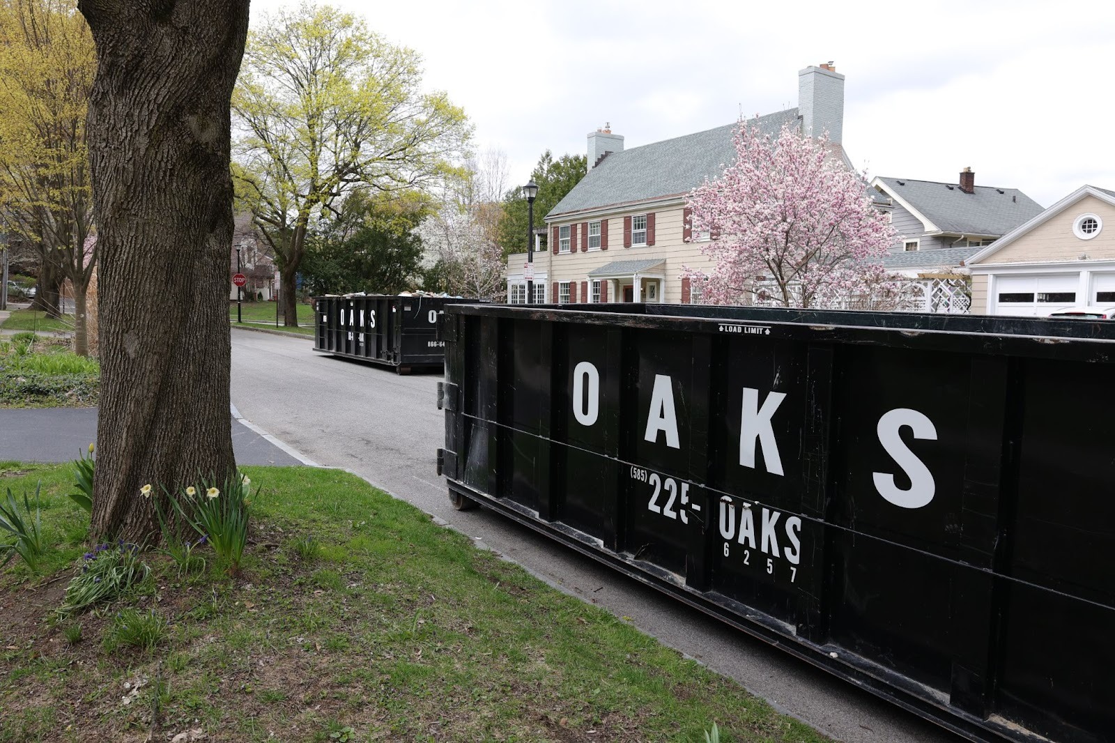 Large black dumpster on a residential street. A house and tree in the background.