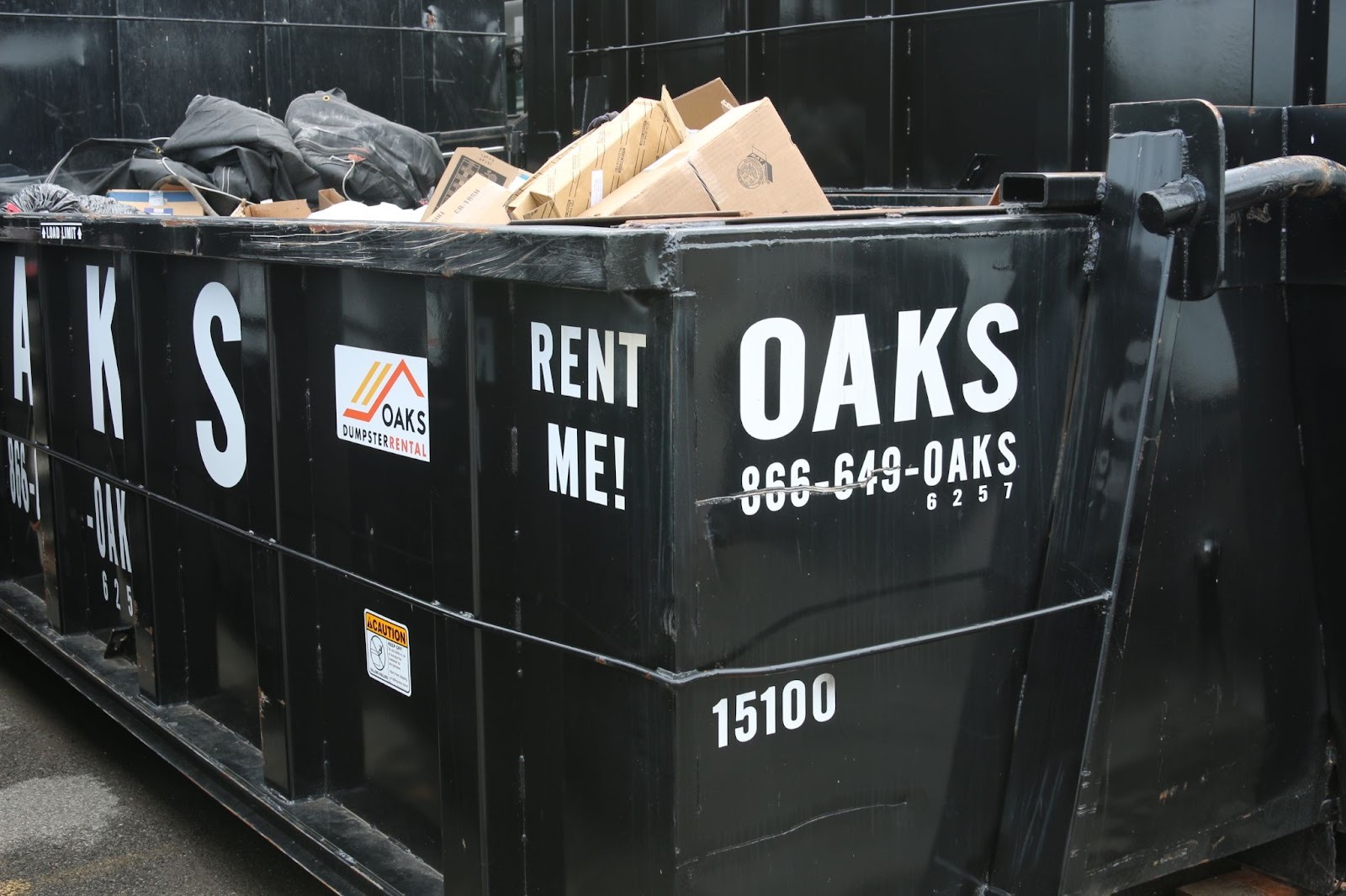 A black dumpster with Oaks and Rent Me text. The dumpster is filled with trash.