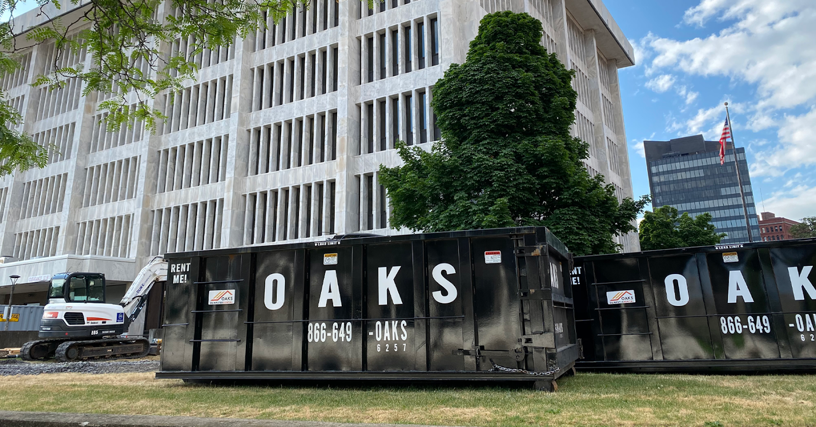A large black dumpster with 'OAKS' written on it sits on a grassy area. The dumpster is likely for waste management.