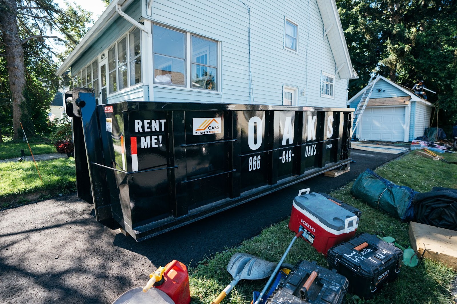 Large black dumpster on residential driveway. Various tools and equipment nearby.