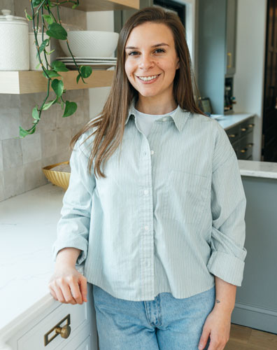 A woman with long brown hair wearing a light blue button-down shirt and jeans. She is standing in a kitchen with a plant and shelves in the background.