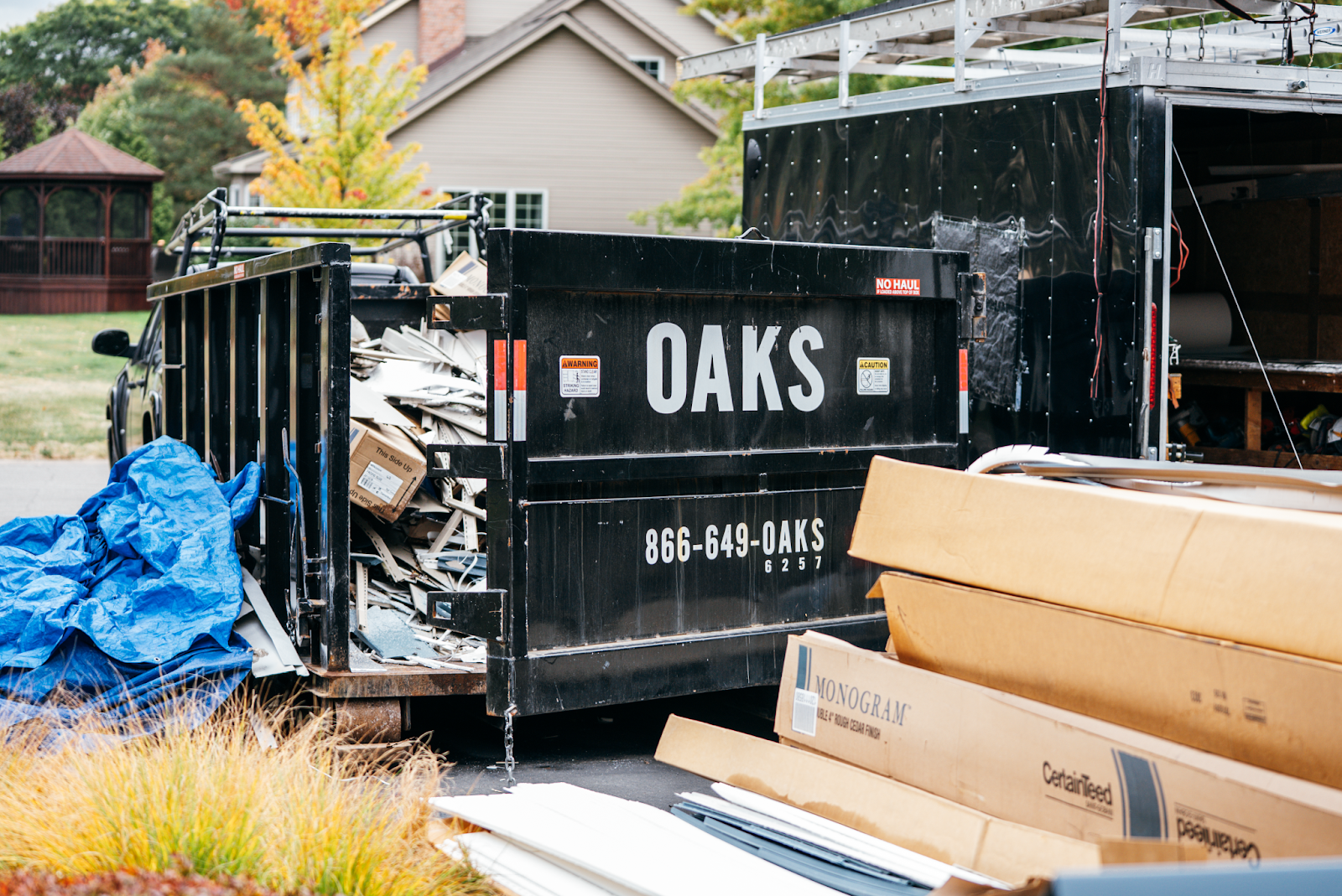 A black dumpster with Oaks logo and phone number. Construction materials are piled next to it.