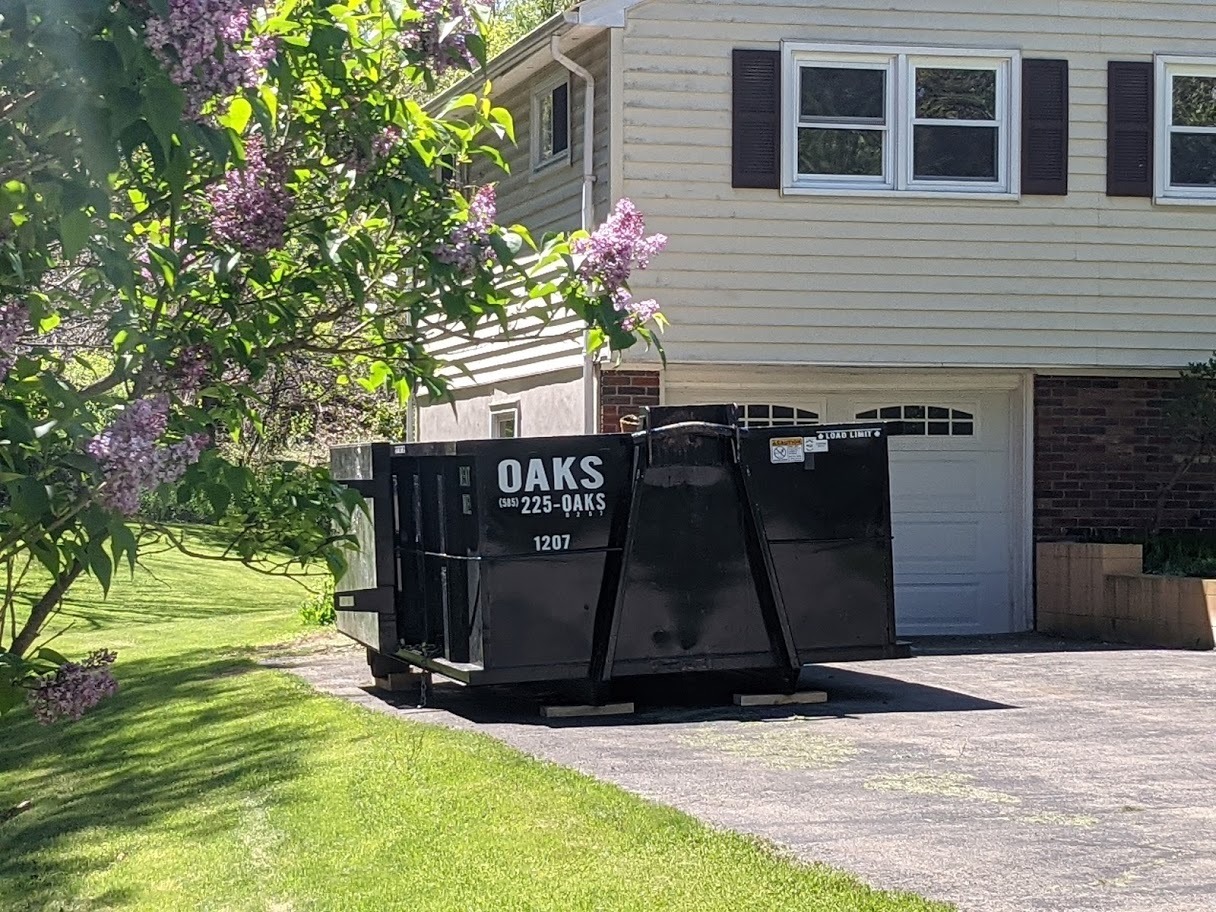 A large black dumpster on a driveway. Oaks Disposal dumpster.