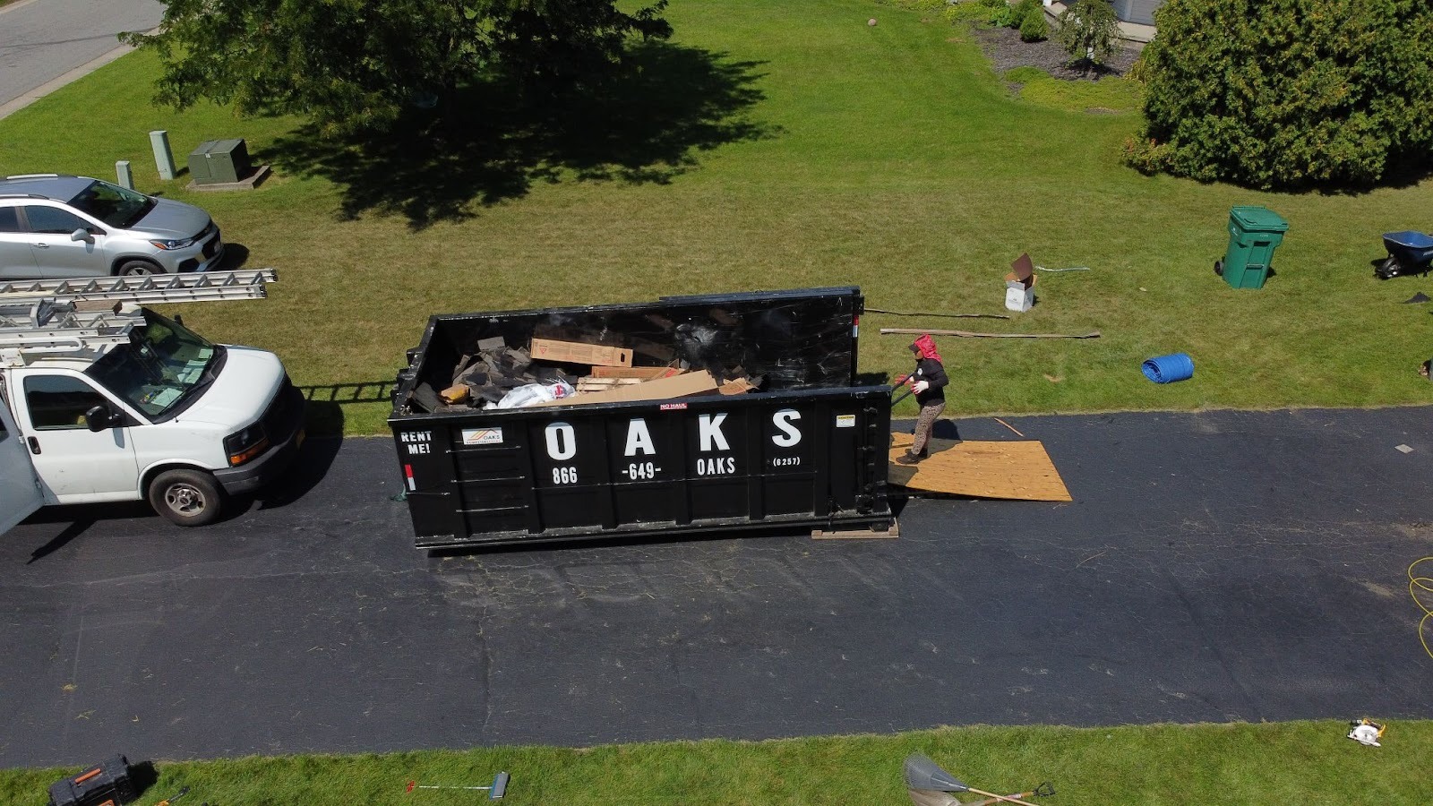 A white truck is parked next to a large black dumpster on a residential street. The dumpster has 'OAKS' written on the side. A person in a red shirt is standing nearby. The scene is set in a quiet neighborhood with grassy areas and trees.
