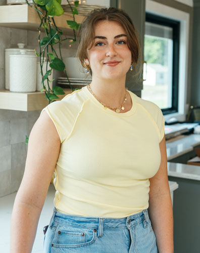 A young woman stands in a kitchen with a plant and jars on a shelf behind her. She is smiling and wearing a yellow shirt and blue jeans.