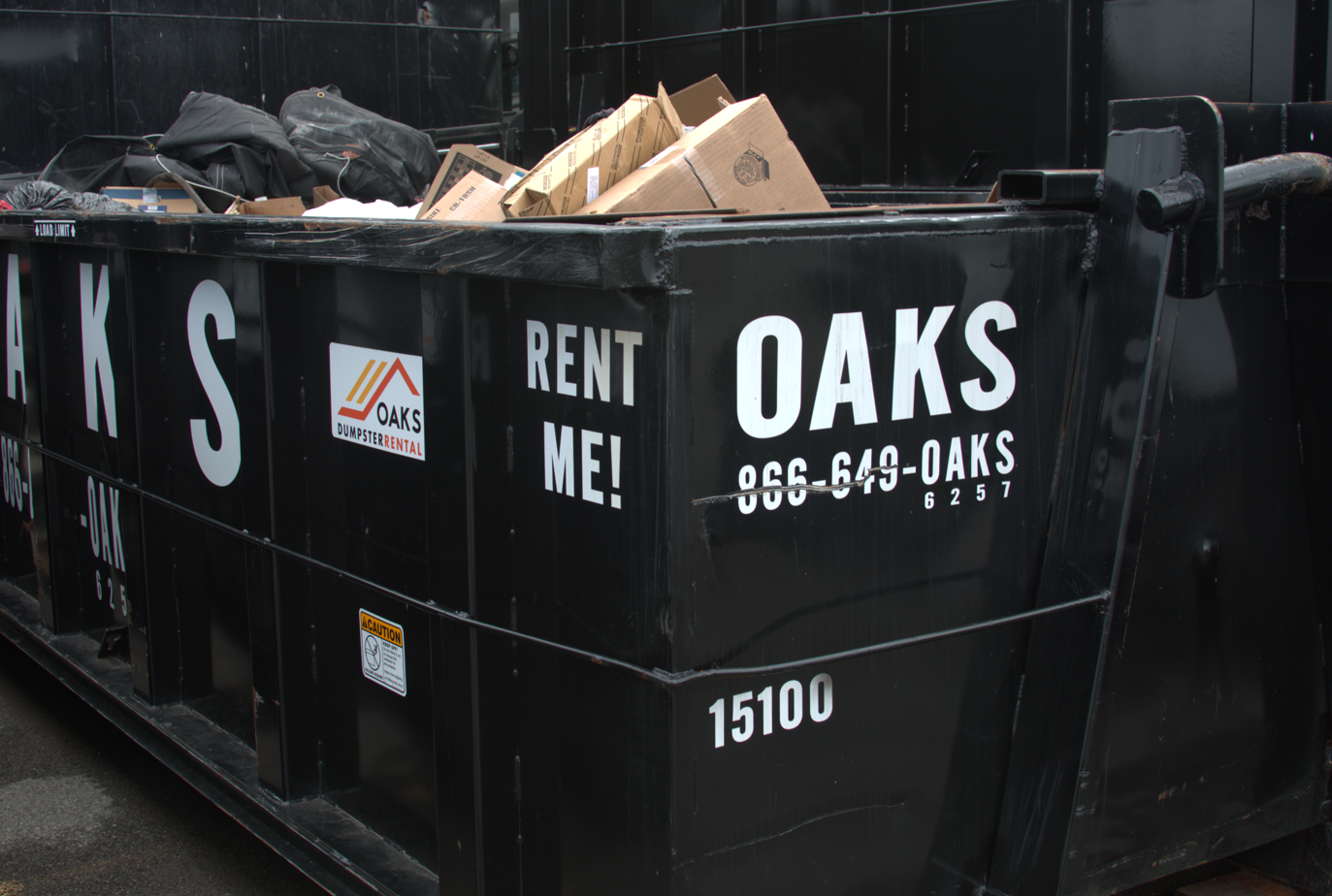 A black dumpster with 'OAKS' and 'RENT ME!' text. Construction waste inside.
