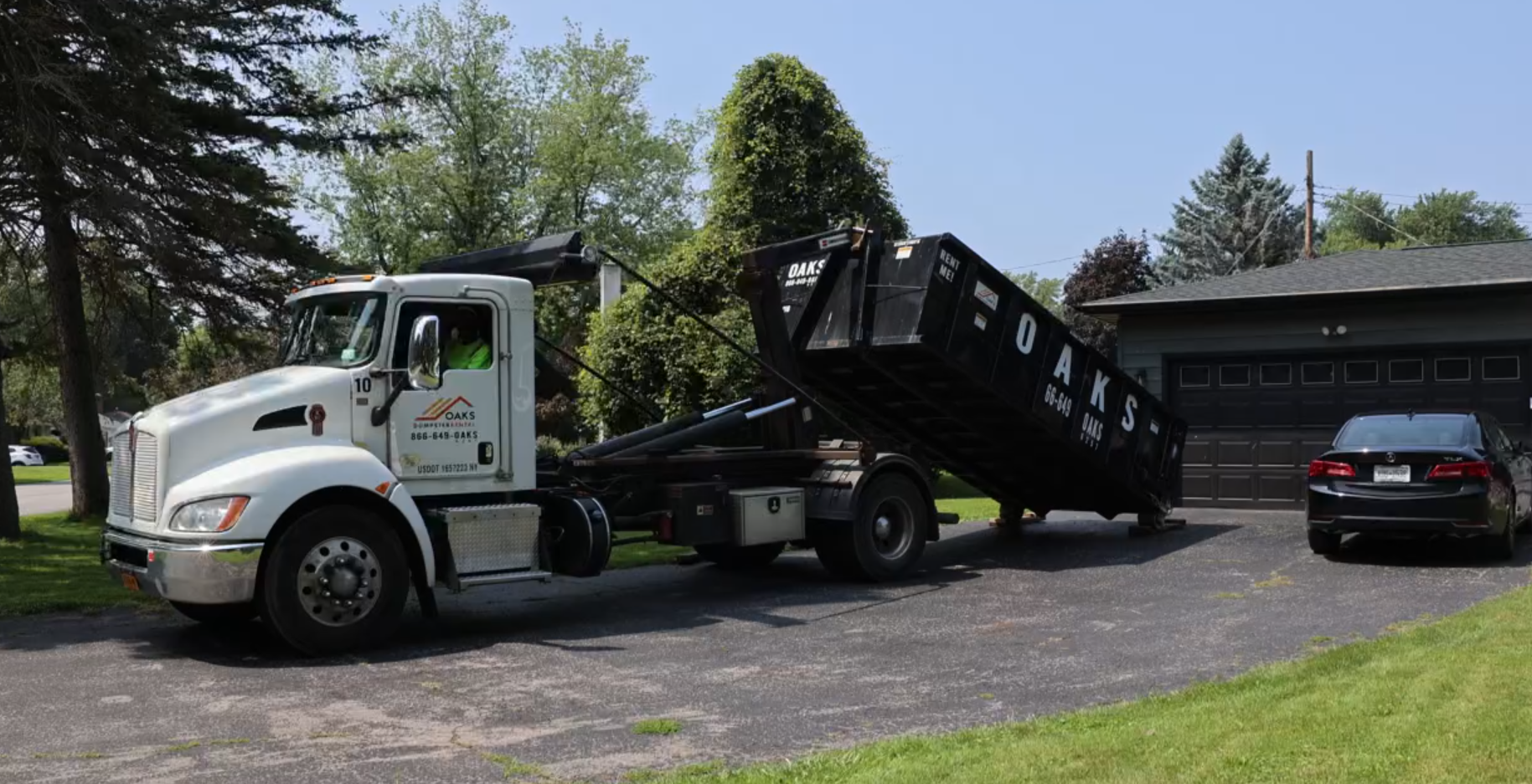 A white truck hauls a large black dumpster on a residential driveway.