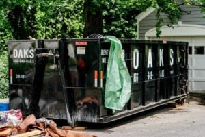A large black container loaded with roof materials, placed by a home. The dumpster features a green liner covering internal waste.