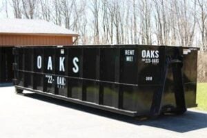 Large black dumpster with 'OAKS' branding for rent. View from driveway during winter season.