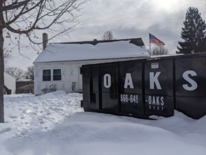 A black dumpster labeled with company information sits in the snow in front of a white building, with a bare tree and evergreens nearby.
