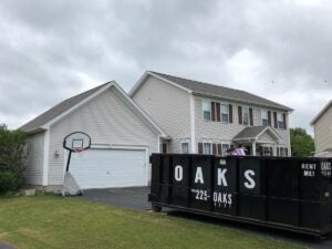 Dark gray two-story house with basketball hoop, driveway, and OAKS dumpster under cloudy skies.