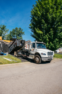 A white dump truck is parked on a paved driveway, hauling a load of rocky fill material in its open bed. The truck is positioned near a grassy area and trees under a bright blue sky.