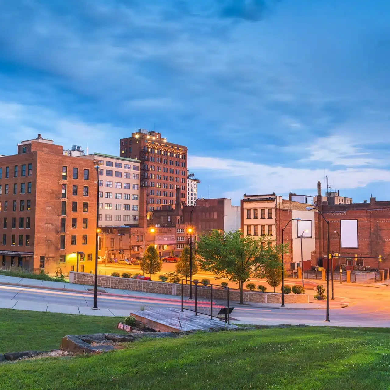 A city street with tall buildings and a grassy area. The scene is lit up in the evening.