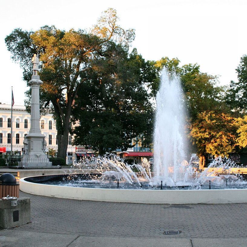 A large fountain in a city park surrounded by trees and a statue.