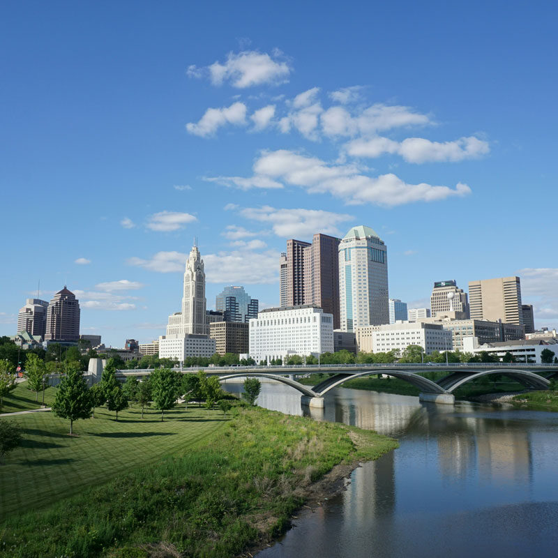 City skyline with a river and bridge on a sunny day. Modern buildings and green spaces.