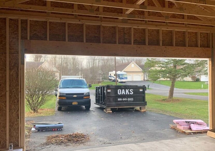 A black dumpster and a white truck parked outside a construction site. Various construction materials are scattered around.