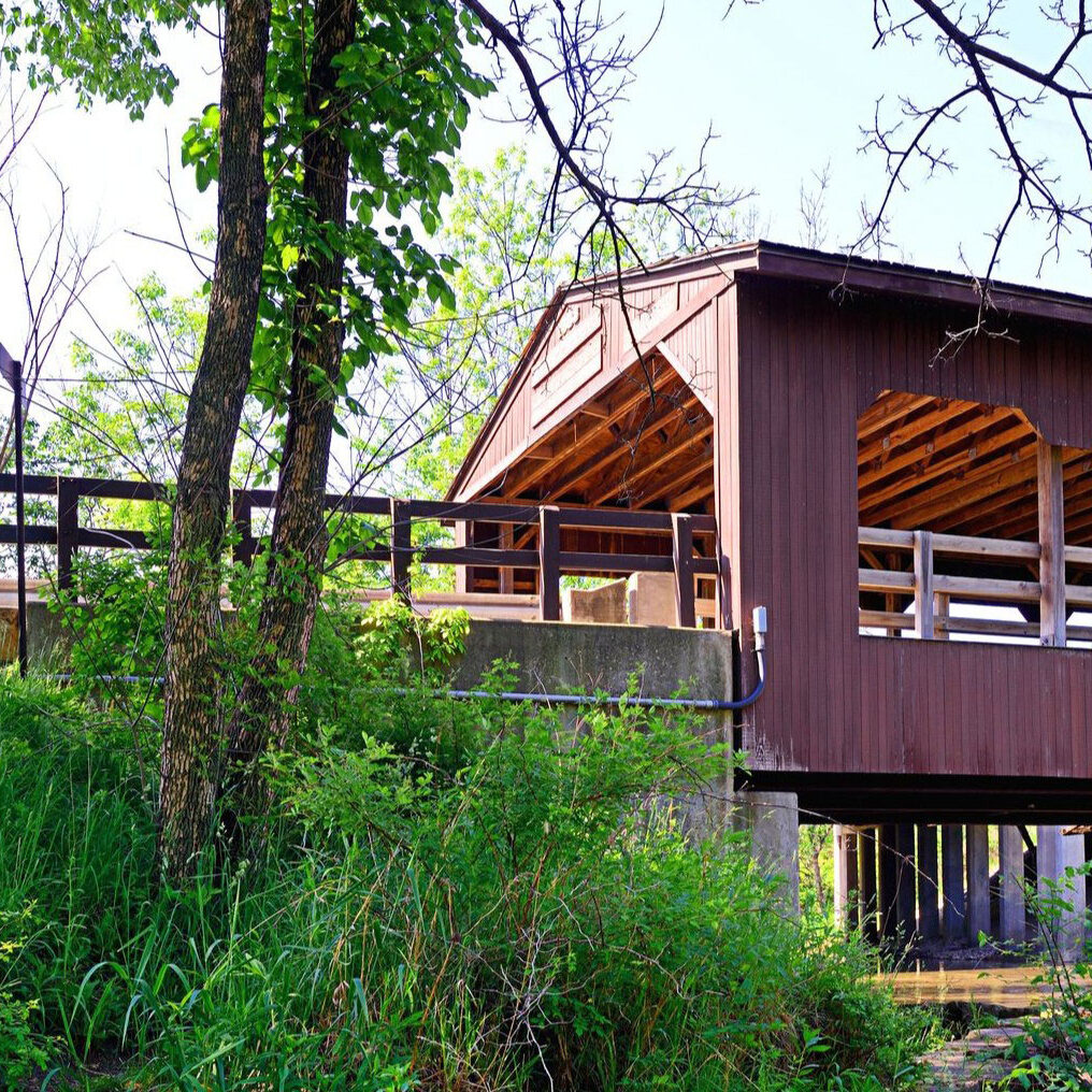 A brown wooden covered bridge surrounded by trees and bushes