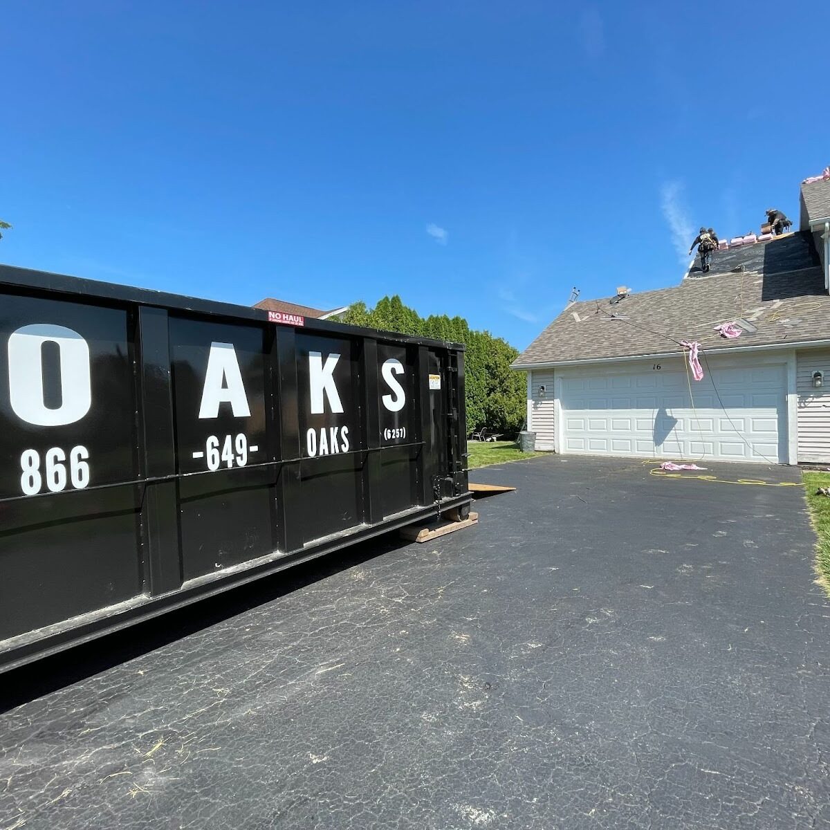 a large black dumpster on a driveway. The dumpster has O-A-K-S and a phone number on it.