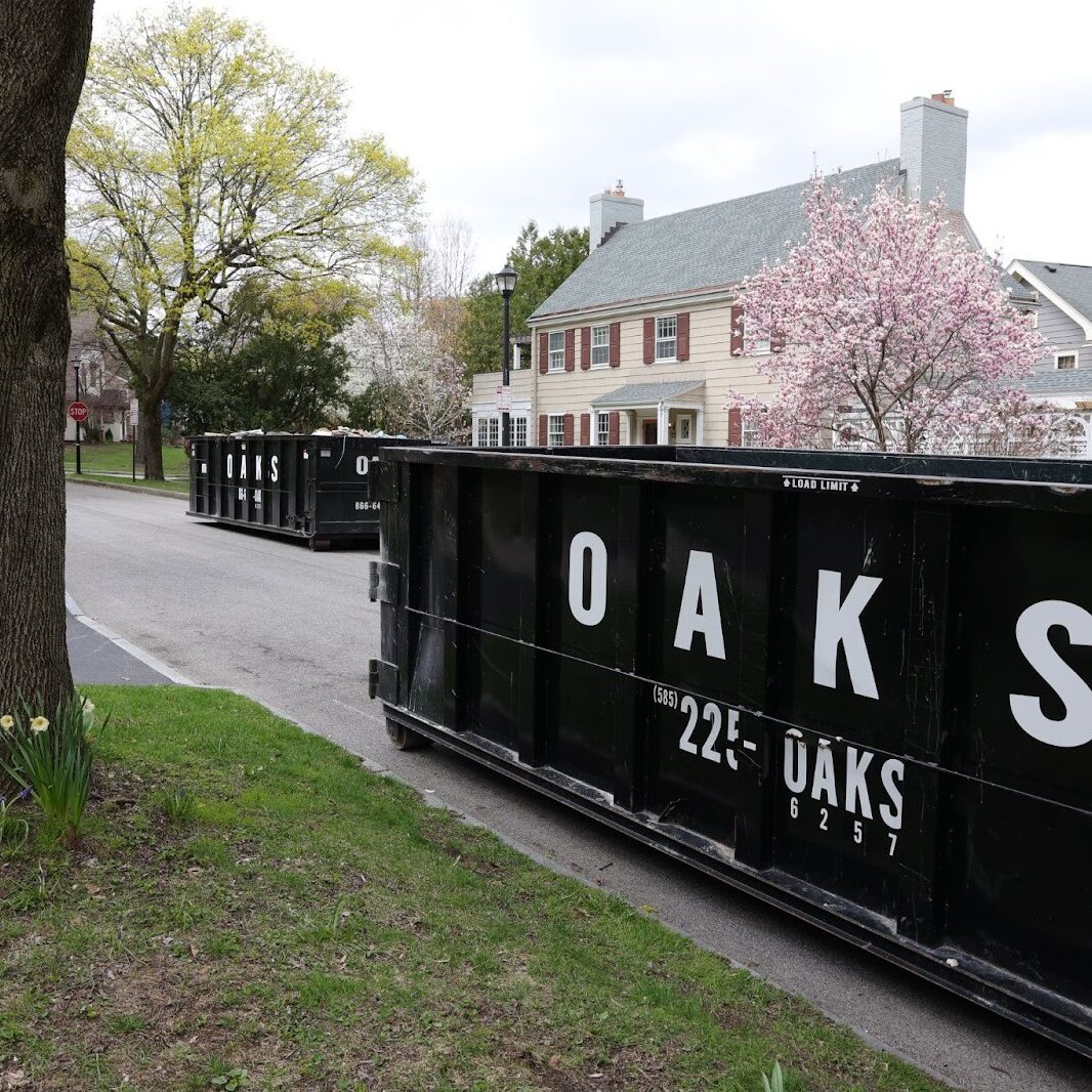 Large black dumpster on a residential street. A house and tree in the background.
