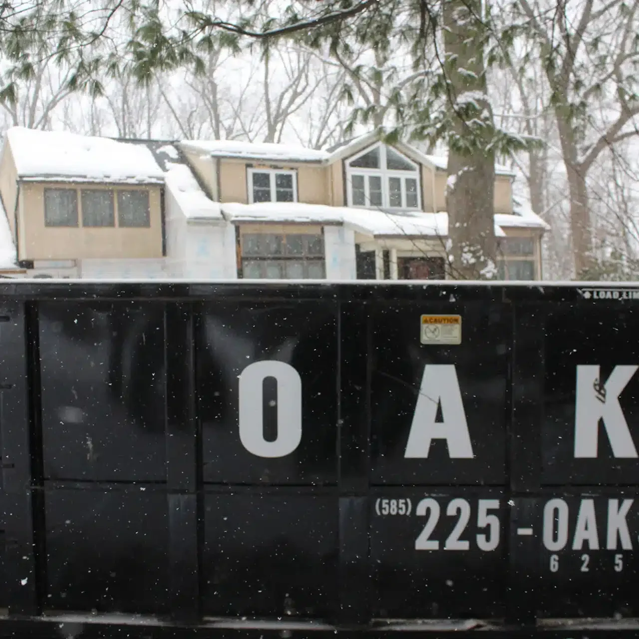 A large black dumpster with white lettering in front of a house covered in snow.