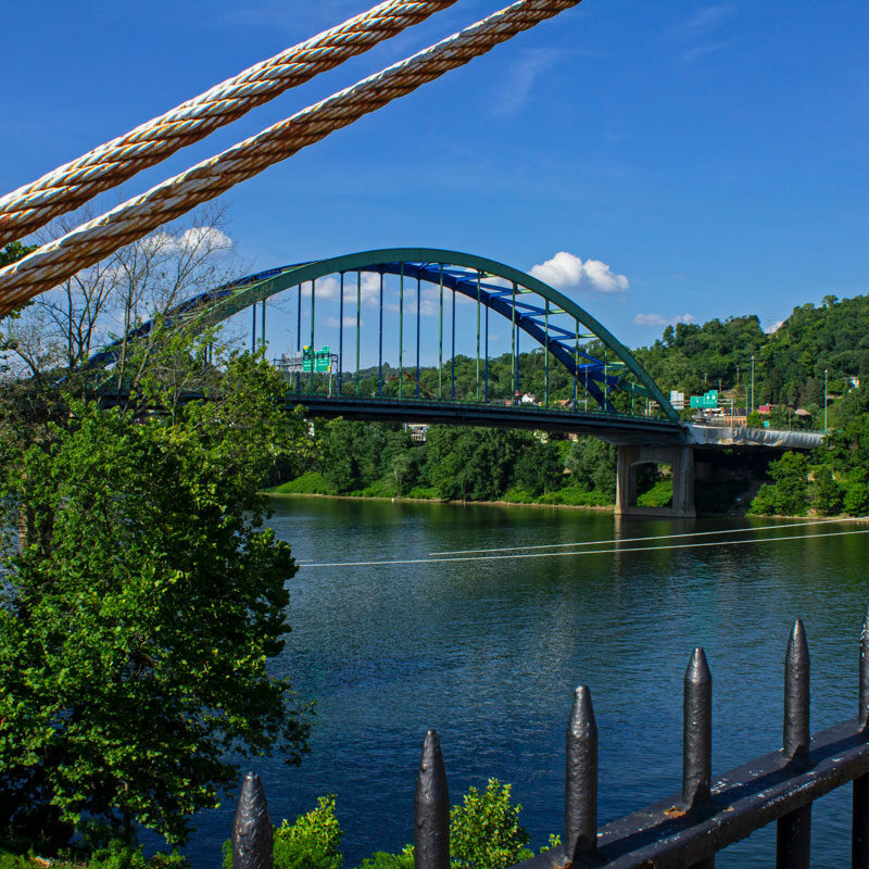 A green arch bridge spanning a river with lush greenery on both sides. A fence in the foreground.