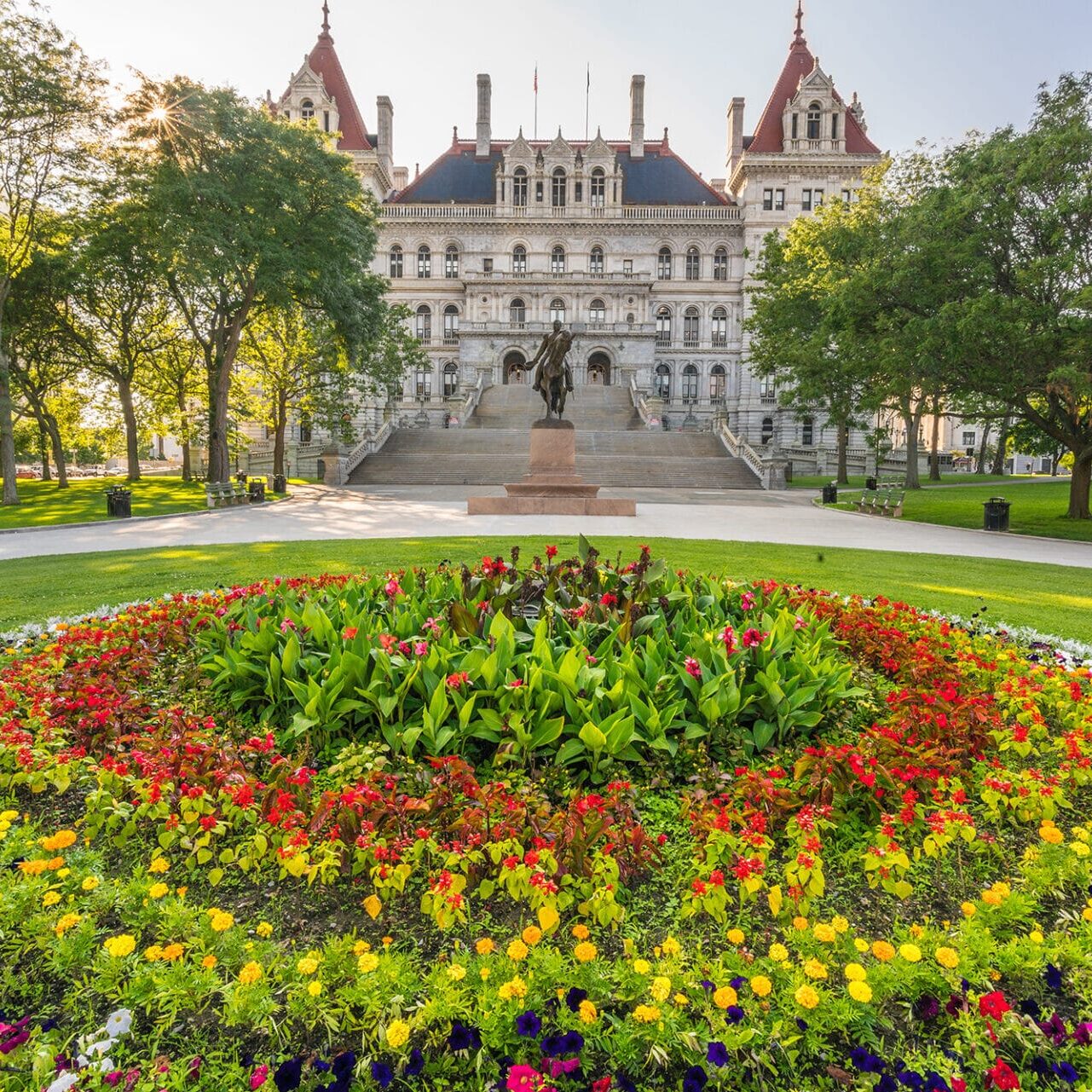 A vibrant flower bed in a park with a historic building and statue in the background. The building features a grand staircase and ornate architecture.