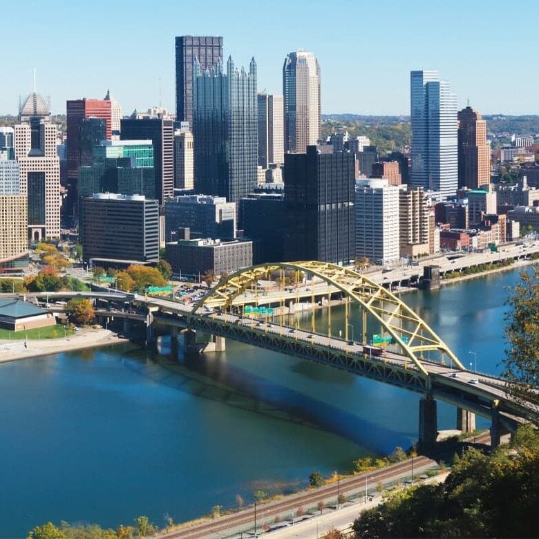 Pittsburgh city skyline with a yellow bridge over a river. The bridge connects the city to a park.